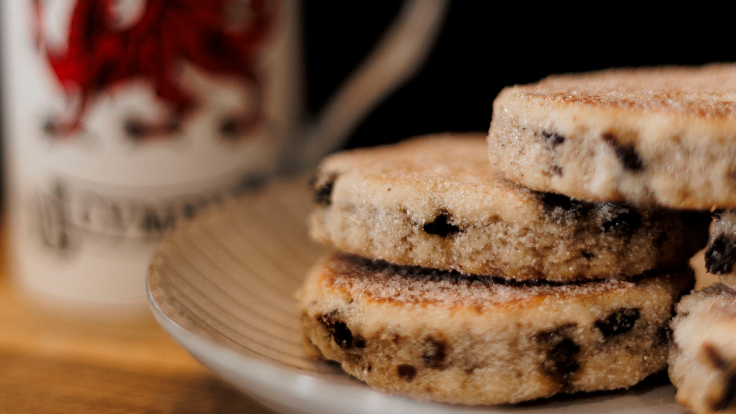 Stack of Welsh Cakes on a plate with a blurred mug  in the background showing a Welsh dragon.